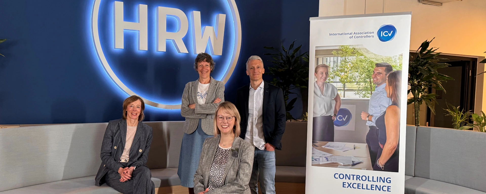 Fünf Personen posieren in einem modern eingerichteten Innenraum vor einer großen blau leuchtenden HRW-Wand. Rechts steht ein Roll-up-Banner der „International Association of Controllers (ICV)“ mit dem Slogan „Controlling Excellence“. Zwei Frauen sitzen vorne, eine weitere Frau und ein Mann stehen dahinter. Die Gruppe wirkt freundlich und professionell. Im Hintergrund sind Pflanzen und Sitzbänke zu sehen.
