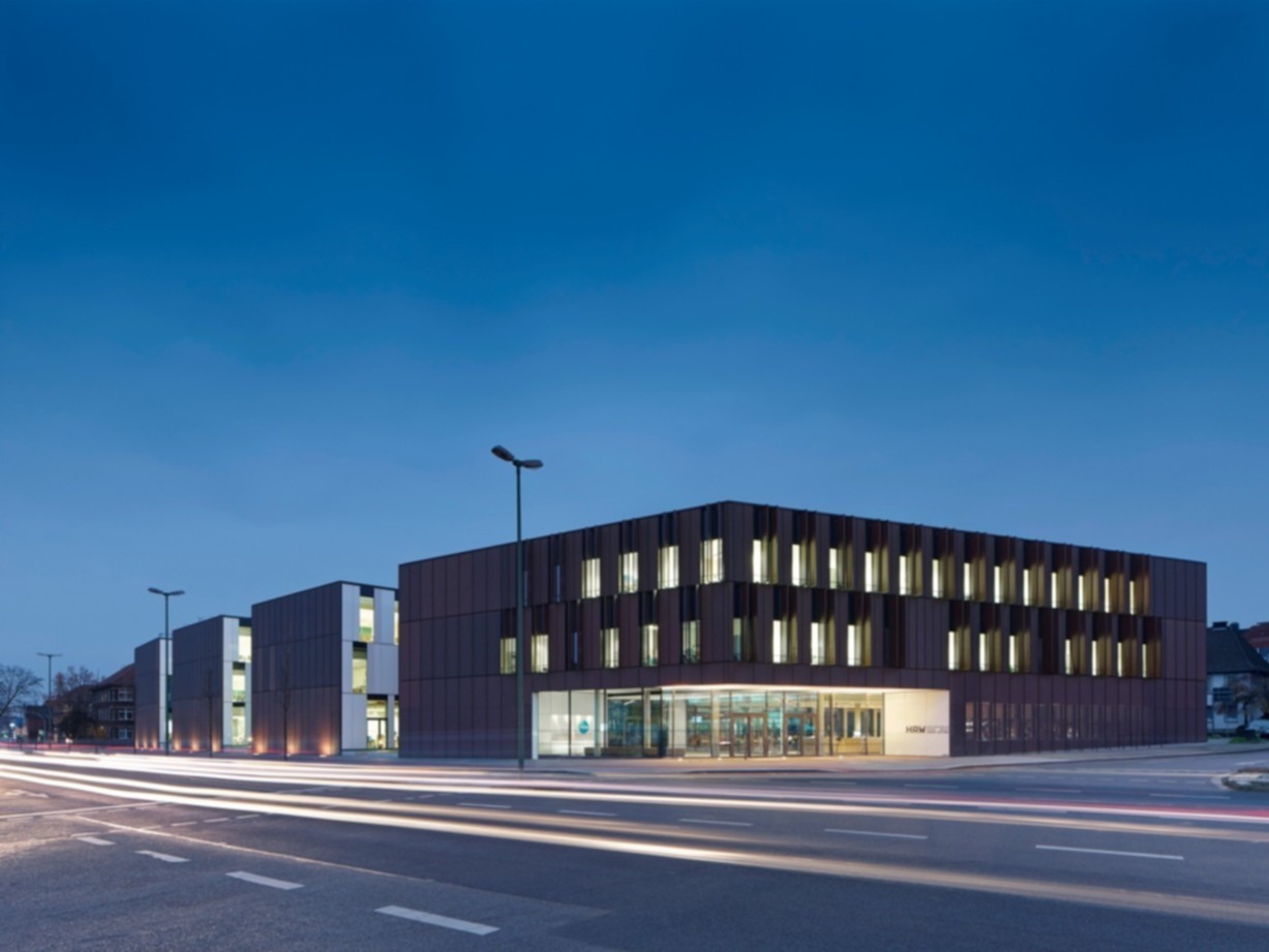 Modernes, mehrstöckiges Hochschulgebäude mit großen Glasfronten und schmalen, vertikalen Fenstern, abends von innen beleuchtet.