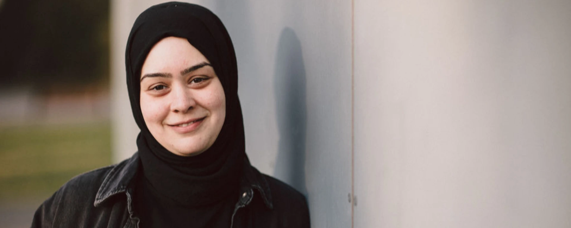 Eine Frau mit schwarzem Kopftuch lehnt an einer Wand.
