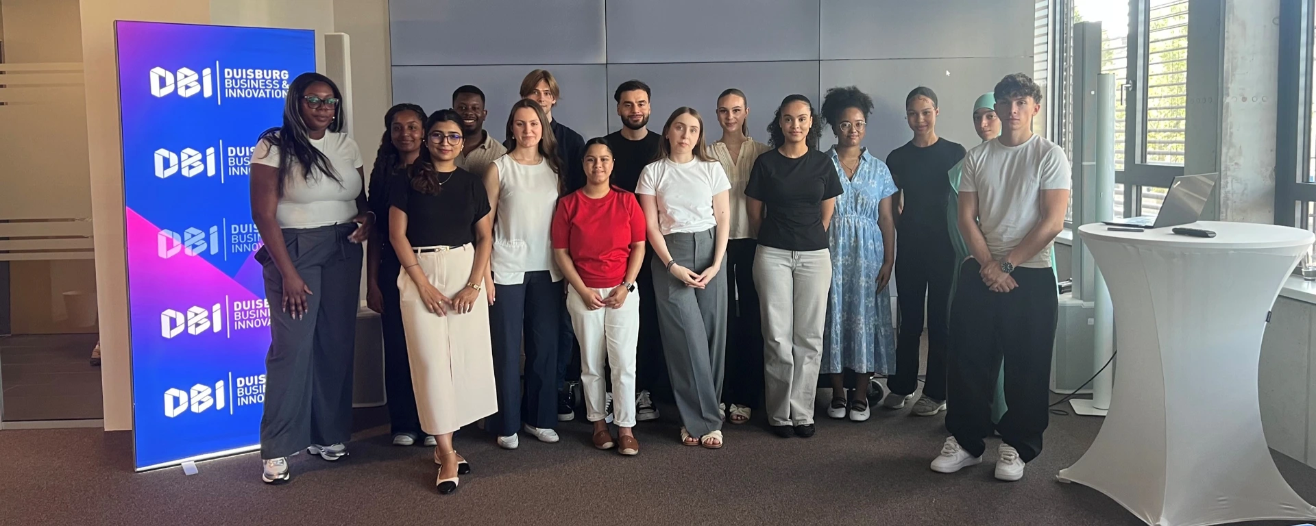 Gruppenfoto von etwa 13 Personen in einem modernen Veranstaltungsraum vor einer großen DBI-Displaywand („Duisburg Business & Innovation“). Die Gruppe steht eng beieinander und lächelt in die Kamera; rechts ist ein Stehtisch mit Laptop zu sehen, im Hintergrund große Fenster mit Tageslicht.