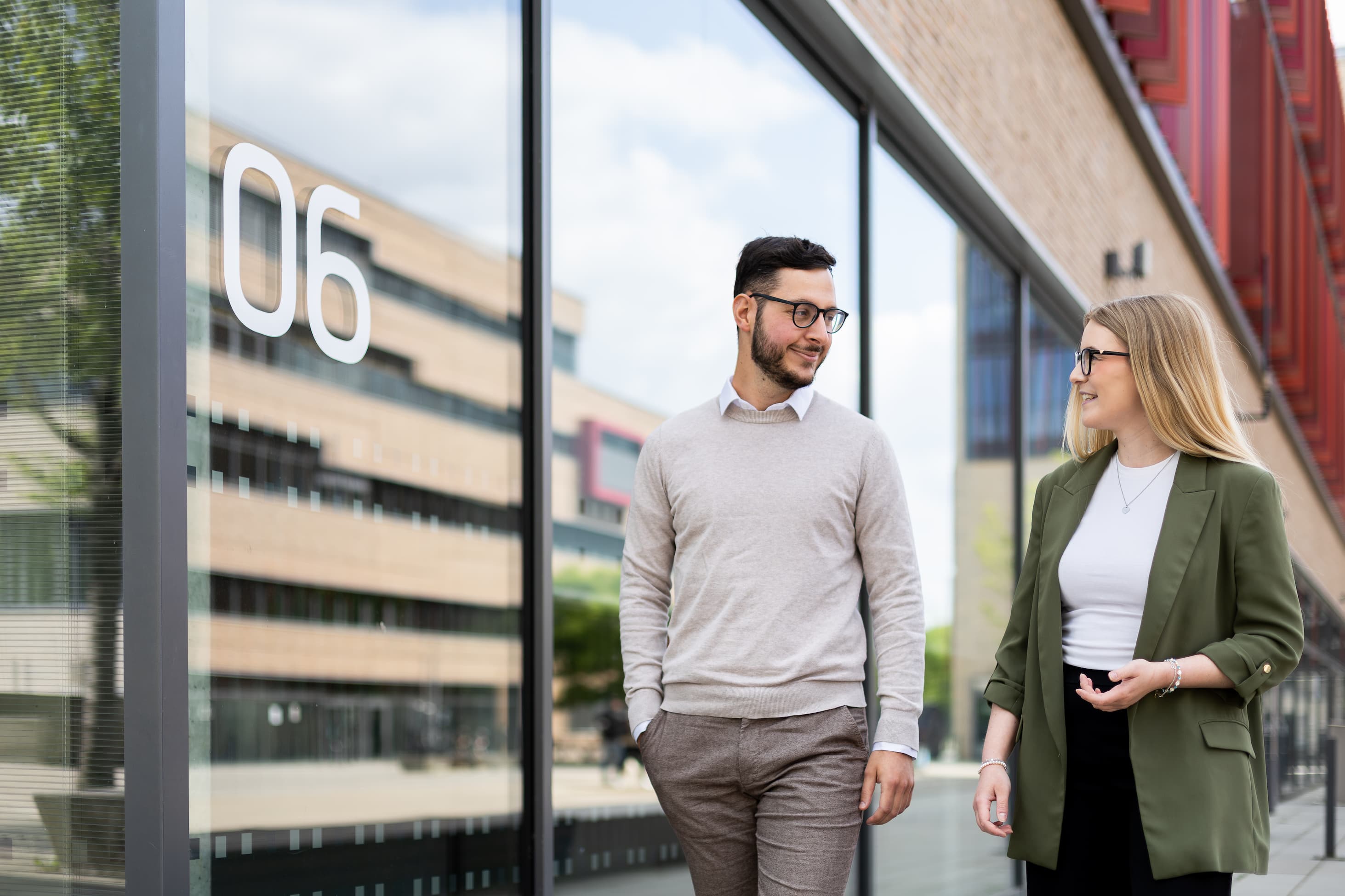 Das Foto zeigt zwei Studierende, die sich auf dem Campus vor dem Hörsaalgebäude unterhalten.