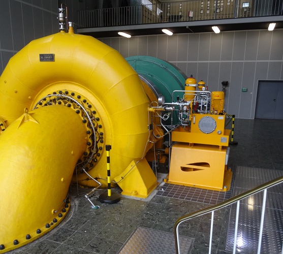 Innenansicht eines Wasserkraftwerks mit einer großen gelben Turbine und zugehöriger Maschinerie. Die Turbine ist mit zahlreichen Bolzen und Flanschen versehen, die ihre Funktionsweise als Teil einer Wasserkraftanlage andeuten. Im Hintergrund sind Schalttafeln und weitere technische Ausrüstungen zu erkennen, die für den Betrieb und die Steuerung der Anlage notwendig sind. Die Anlage wirkt modern und gut instand gehalten, mit einem Fokus auf Effizienz und Energiegewinnung.
