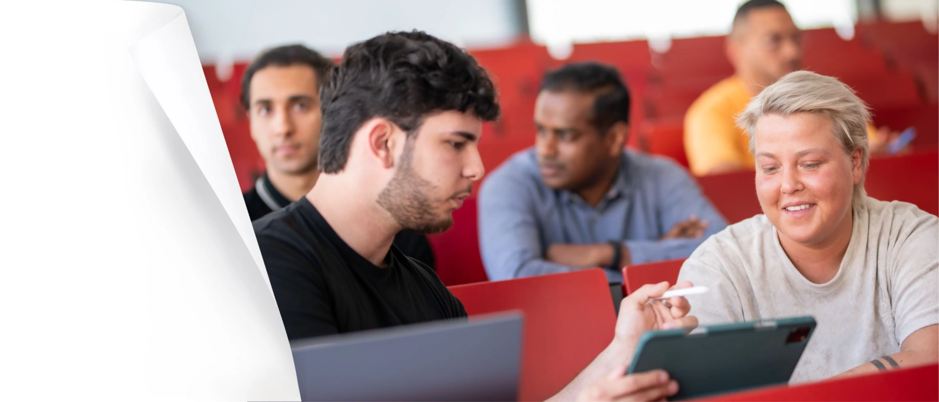 Studierende sitzen in einem Hörsaal mit roten Sitzen; vorn sprechen zwei Personen miteinander und schauen auf ein Tablet, im Hintergrund weitere Studierende unscharf.