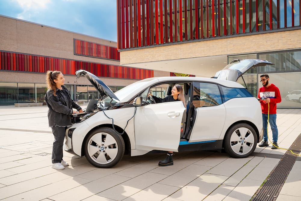 Studierende untersuchen ein Elektroauto auf dem Campus; eine Person sitzt im Fahrzeug, zwei weitere prüfen Motorraum und Kofferraum.