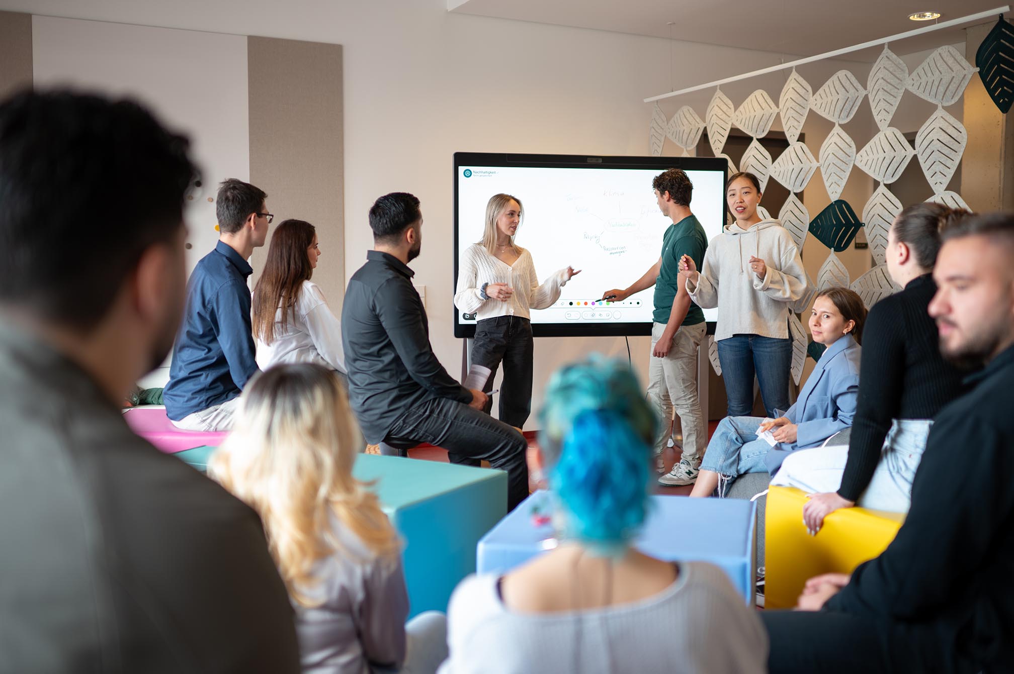 Studierende im Austausch, drei Personen stehen an einem digital Whiteboard, 9 sitzen auf unterschiedlichen, bunten Sitzgelegenheiten dem Whiteboard zugewandt