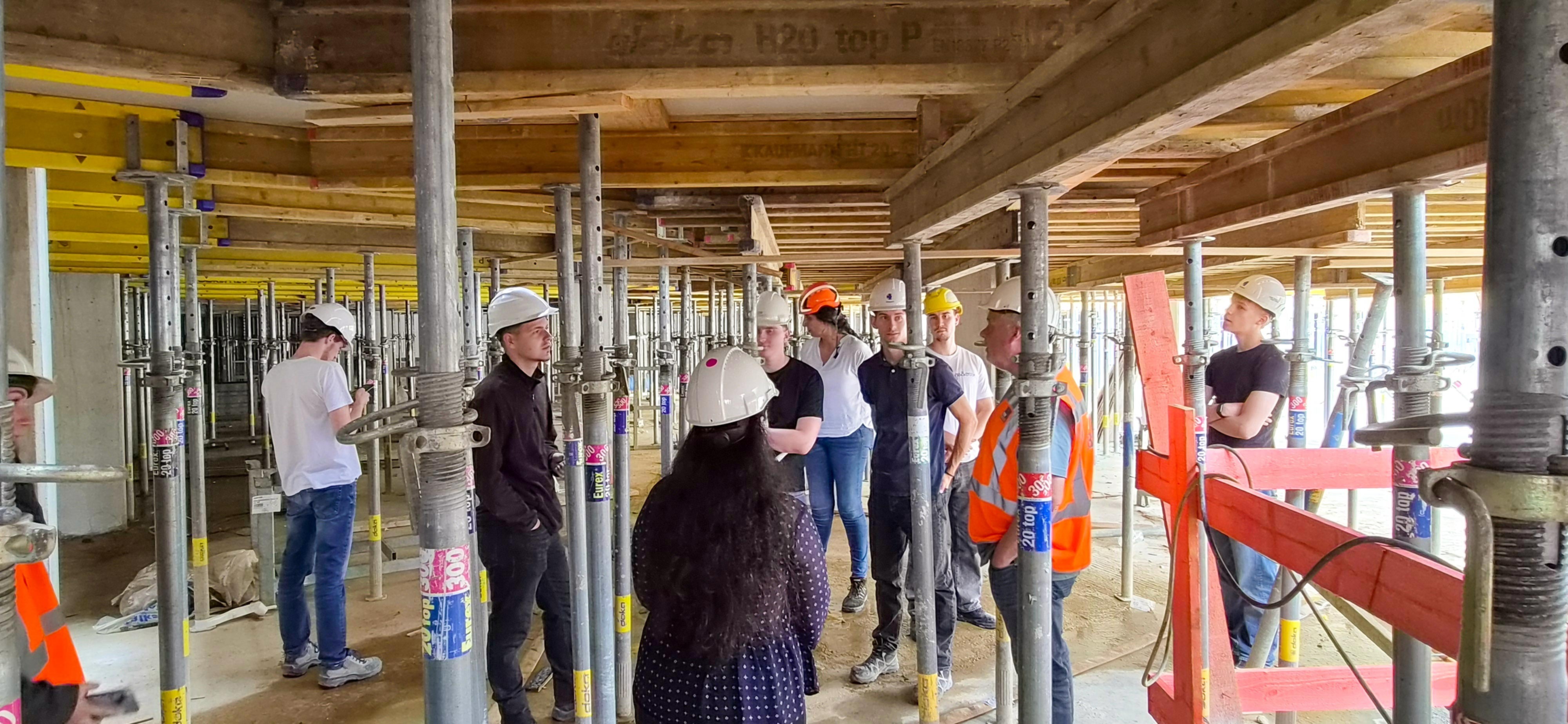 Gruppe von Studierenden mit Schutzhelmen und Warnwesten auf einer Baustelle im Rohbau, umgeben von Deckenstützen und Holzträgern; Besprechung vor Ort.