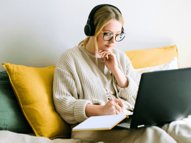 Inmitten farbiger Kissen sitzt eine junge Frau mit Kopfhörern auf den Ohren und Schreibuntensilien vor ihrem Laptop.