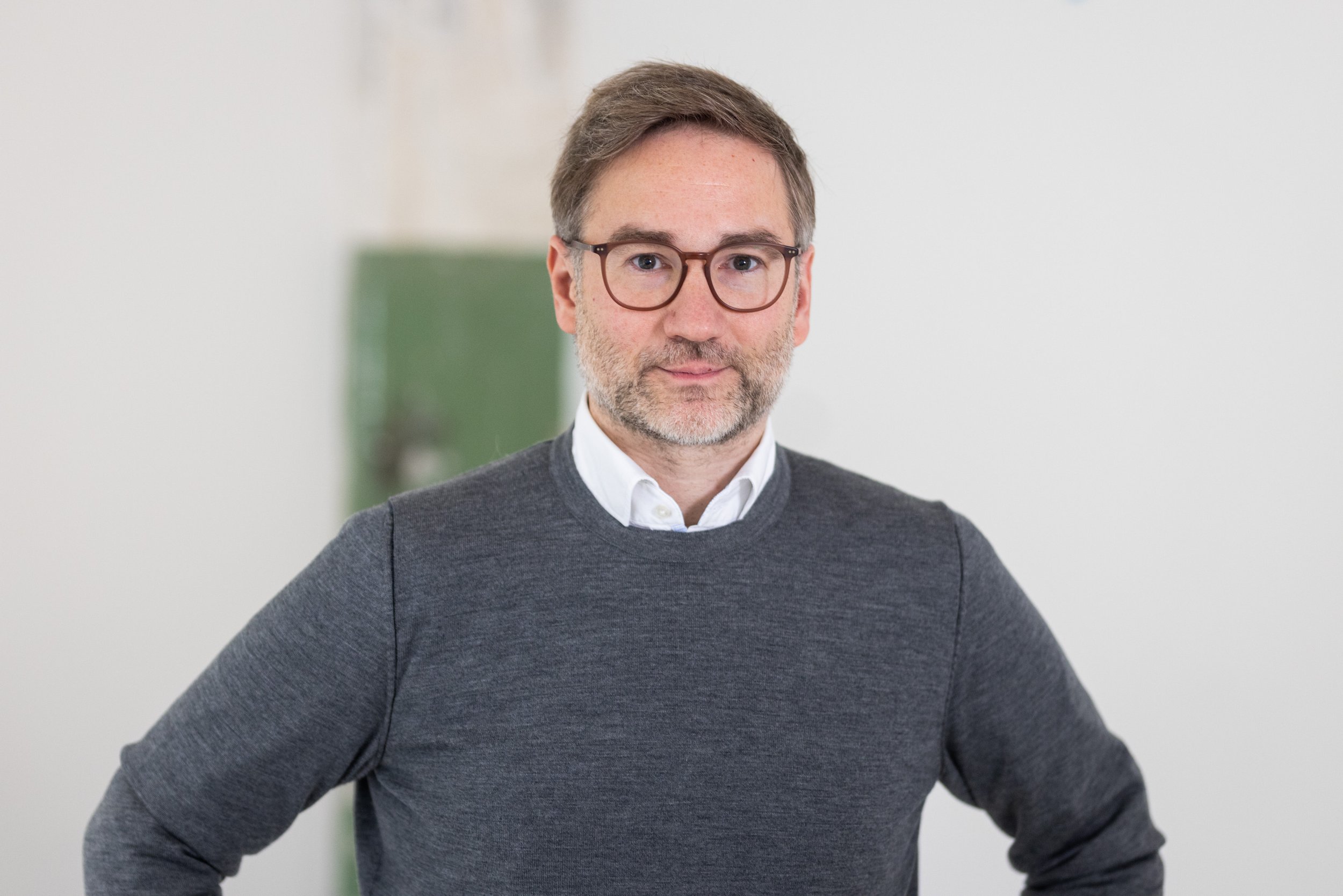 Mann mit Brille und kurzem Bart in grauem Pullover über weißem Hemd, blickt in die Kamera vor heller Wand.