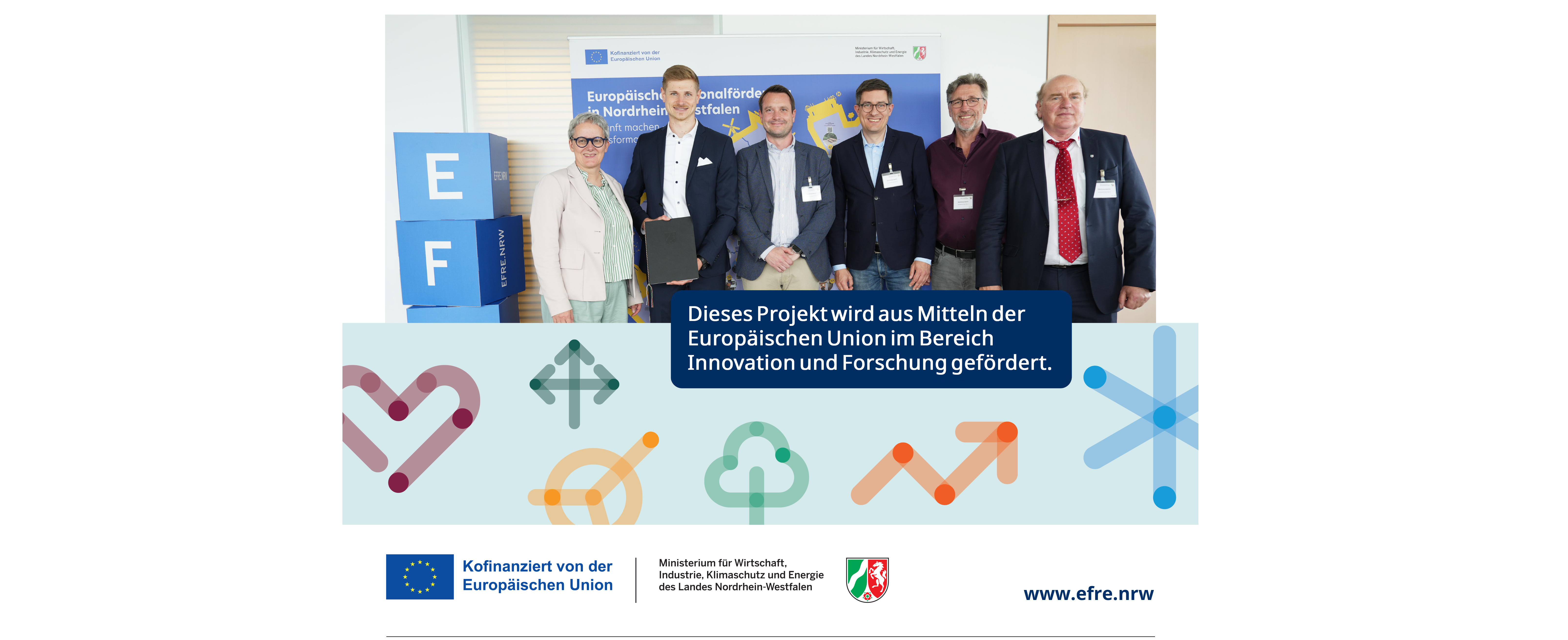 Gruppenfoto von sechs Personen vor einem Roll-Up zur Europäischen Regionalförderung in Nordrhein-Westfalen. Links im Bild drei übereinandergestapelte Würfel mit den Buchstaben ‚E‘, ‚F‘ und ‚R‘. Im Vordergrund ein blauer Kasten mit weißer Schrift: ‚Dieses Projekt wurde aus Mitteln der Europäischen Union im Bereich Innovation und Forschung gefördert.‘ Darunter Symbole in Herz-, Pfeil-, Baum-, Kreis- und Sternform.