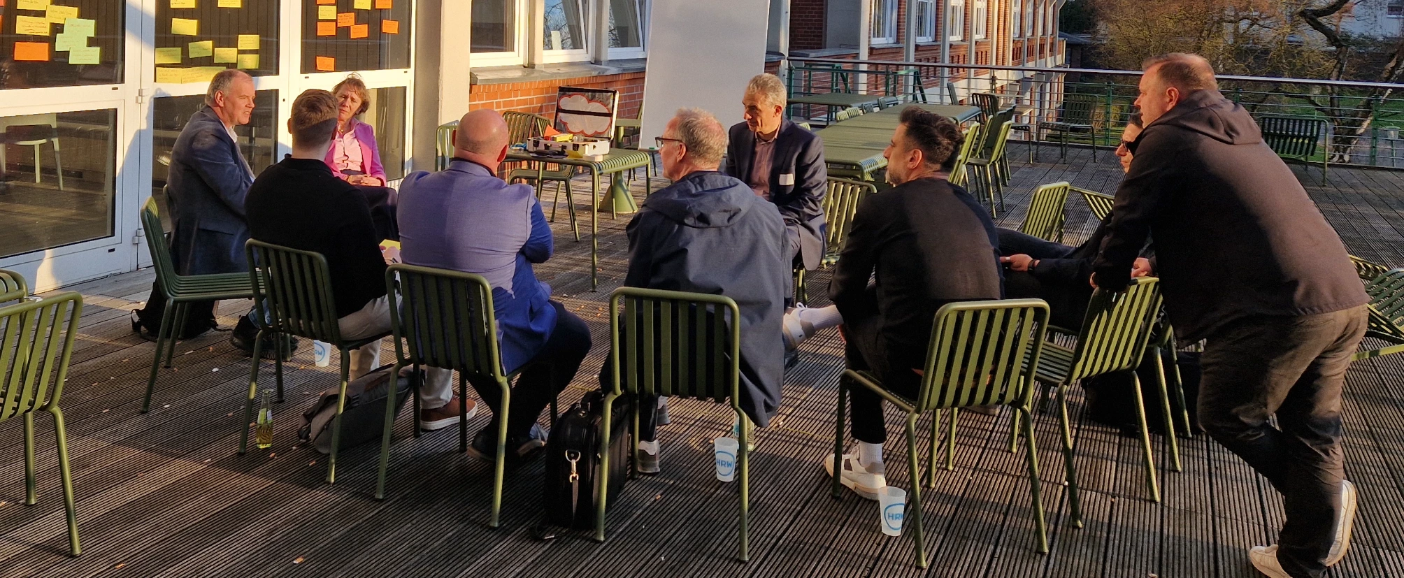 Teilnehmende  sitzen auf einer Terrasse in einem Stuhlkreis und diskutieren gemeinsam; hinter ihnen sind an den Fenstern gesammelte Ideen auf Haftnotizen zu sehen.