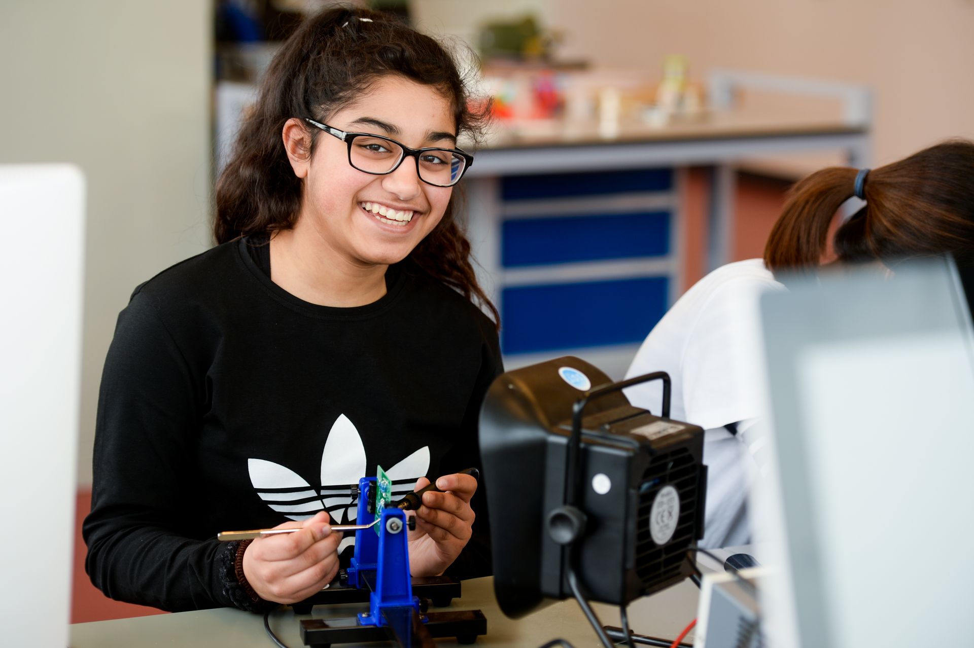 Eine Studentin der Hochschule Ruhr West arbeitet konzentriert an einer Lötstation und lächelt in die Kamera. Das Bild steht für praxisnahes Lernen, MINT-Studiengänge und die Förderung technischer Kompetenzen an der HRW.