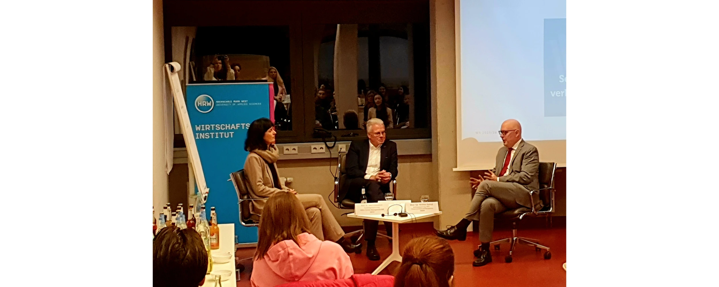 Podiumsdiskusion in einem Seminarraum: Eine Frau und zwei Männer sitzen auf Stühlen im Gespräch vor einem Publikum. Hinter ihnen steht ein blaues Roll-Up mit der Aufschrift ‚Wirtschaftsinstitut‘ sowie eine Leinwand mit Präsentationsfolie. Im Vordergrund sind einige Zuhörerinnen und Zuhörer zu sehen.