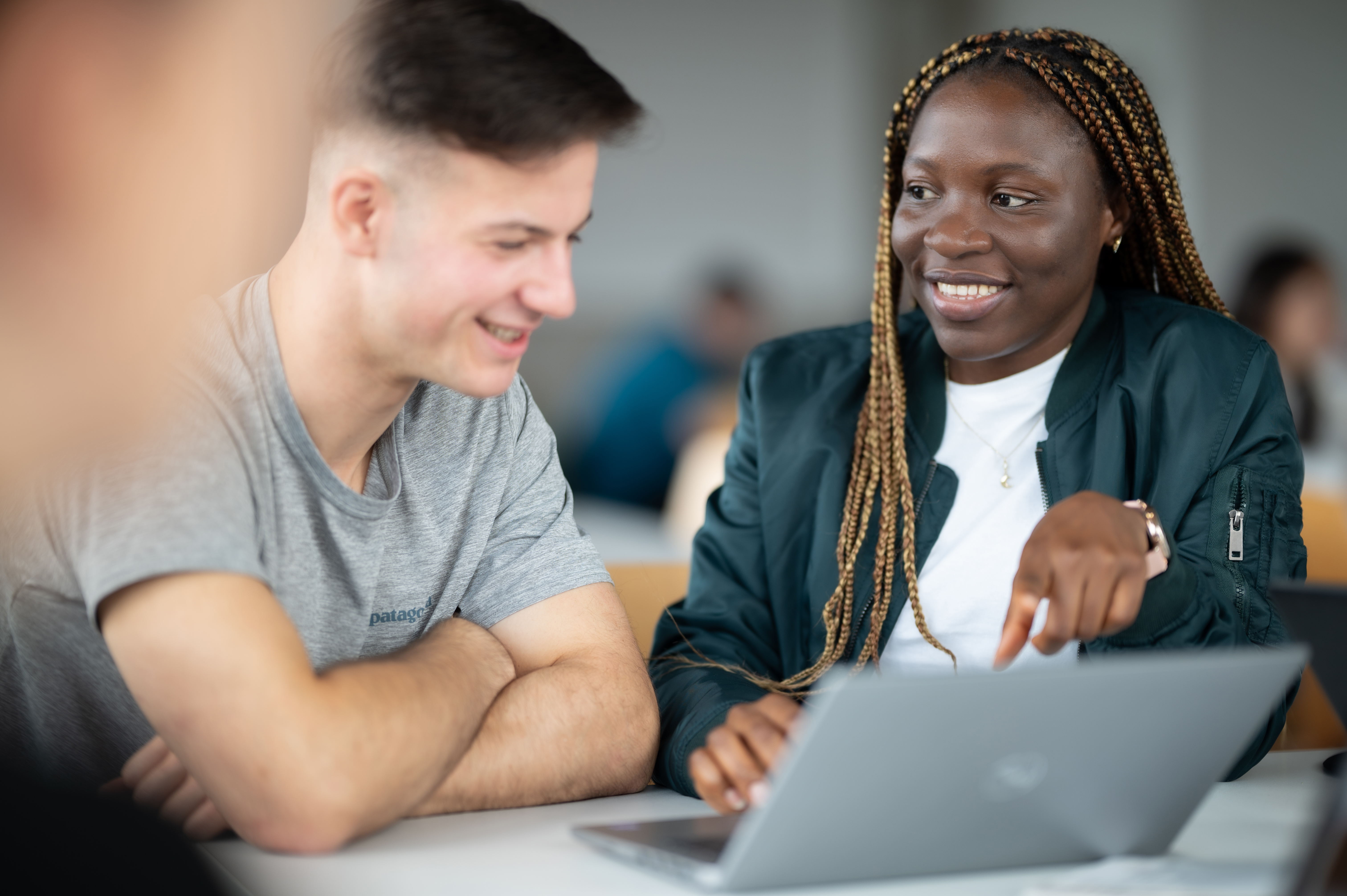 Zwei Studierende sitzen gemeinsam an einem Laptop. Eine Person zeigt auf den Bildschirm, beide lachen und wirken konzentriert und gut gelaunt beim gemeinsamen Arbeiten.