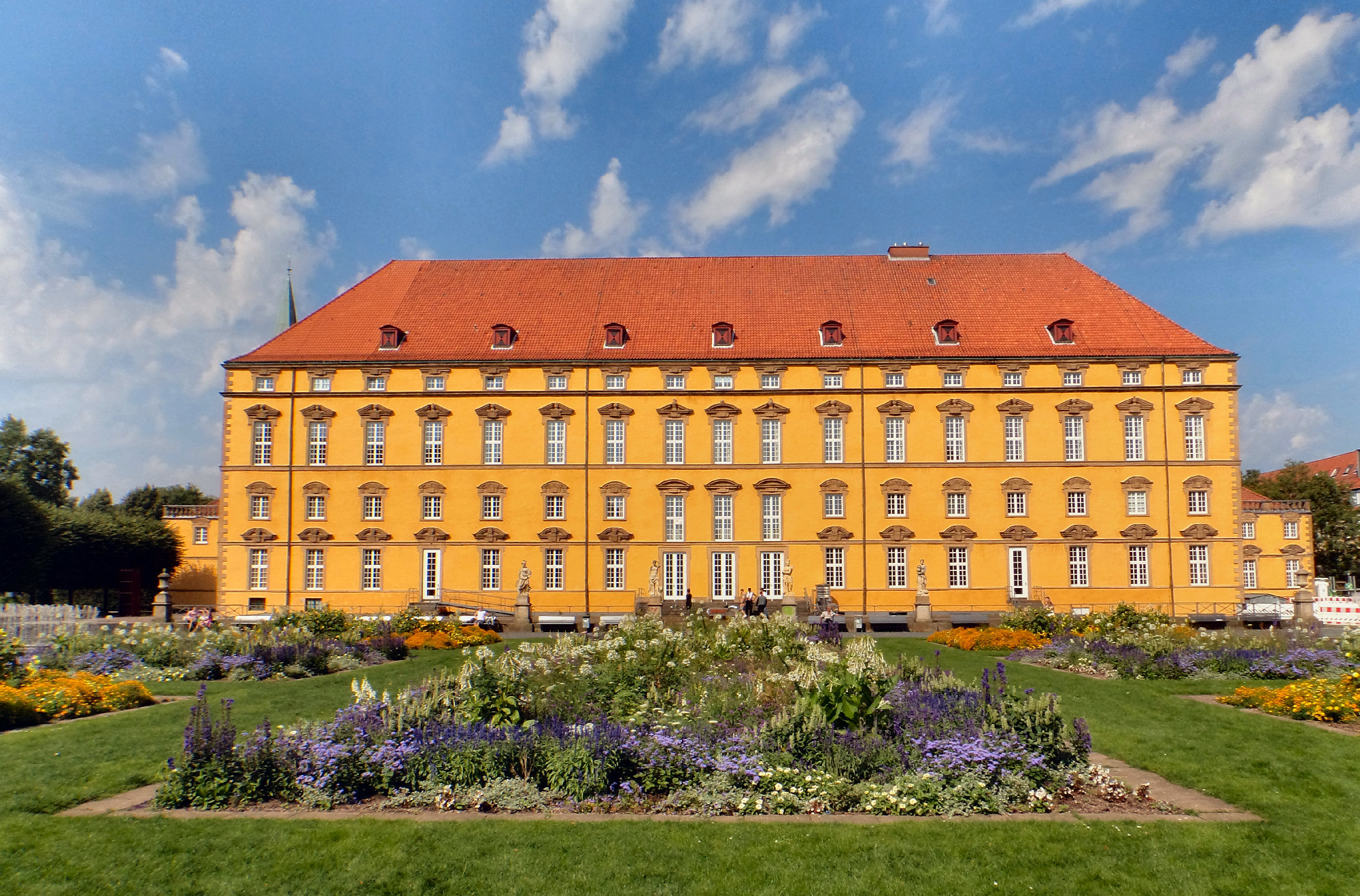 Foto Ein gelber, historsicher Schlossbau mit vielen Fenstern und einen roten Dach unter blauem Himmel im Hintergrund. Im Vordergrund sieht man  Teile einer Grünanlage.
