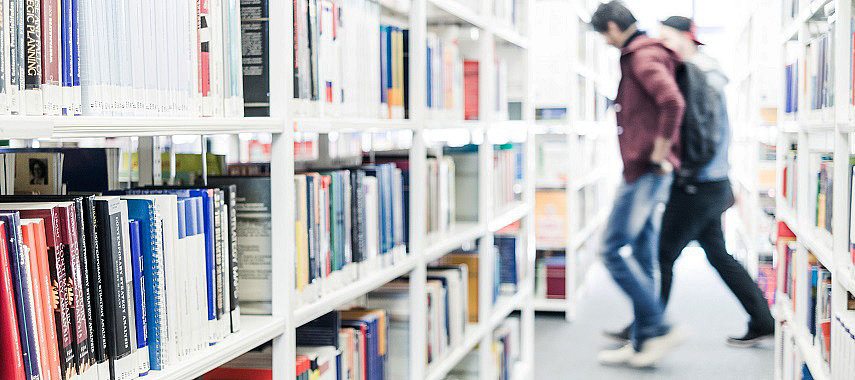Ein Bild von zwei Studenten zwischen Bücherregalen in der Bibliothek