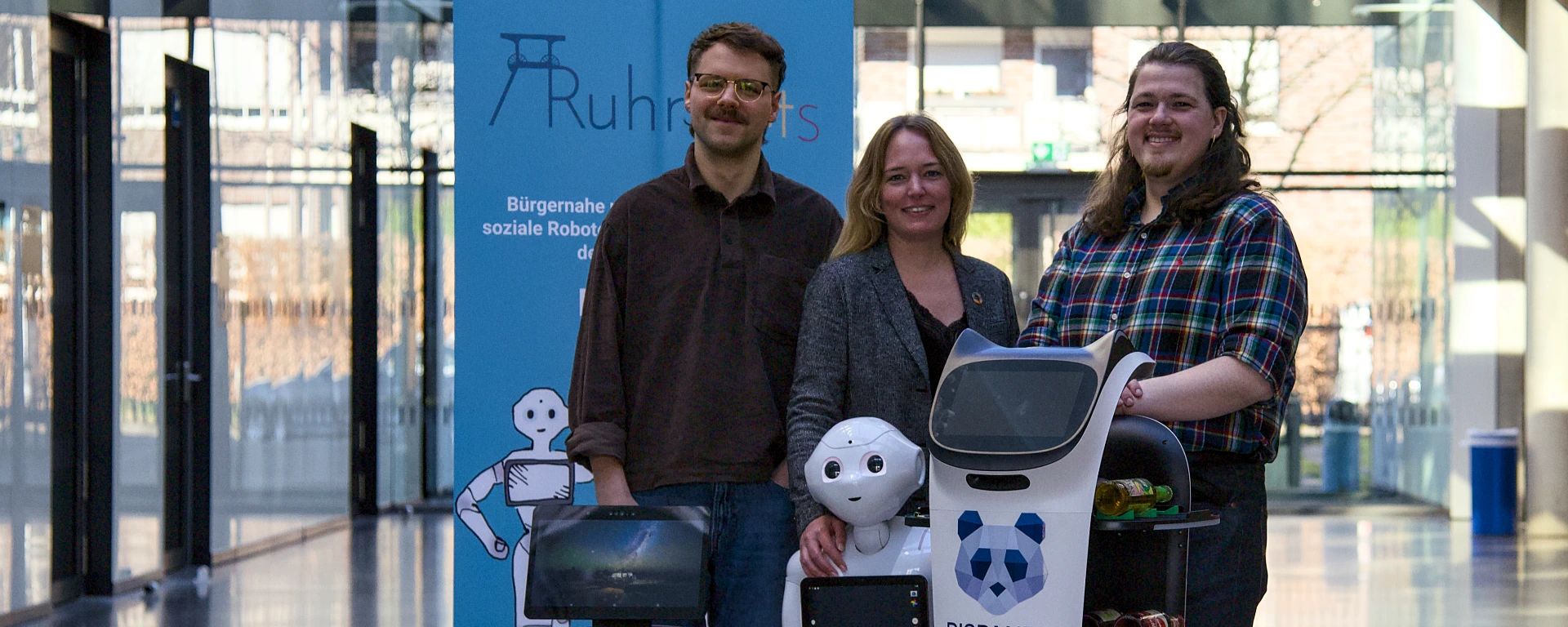 Eine Frau steht zwischen zwei Männern, vor ihnen steht ein humanioder Roboter.