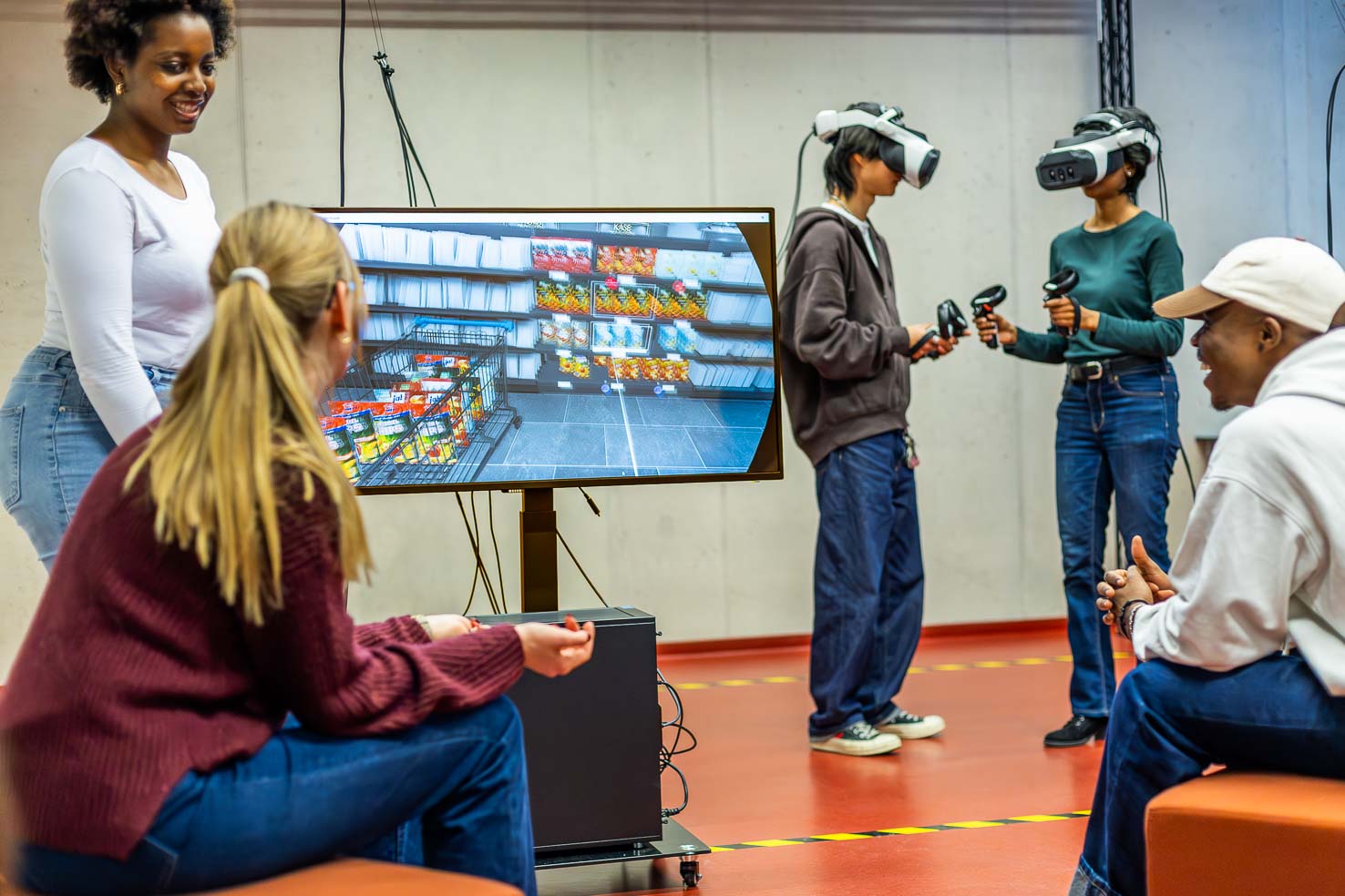 Eine Gruppe junger Erwachsener nimmt an einer Virtual-Reality-Demonstration in einem Innenraum teil. Zwei Personen tragen VR-Headsets und halten Controller in den Händen, während sie neben einem großen Bildschirm stehen, auf dem eine Szene aus einem Supermarkt zu sehen ist. Drei weitere Personen sitzen oder stehen in der Nähe, beobachten das Geschehen und unterhalten sich.