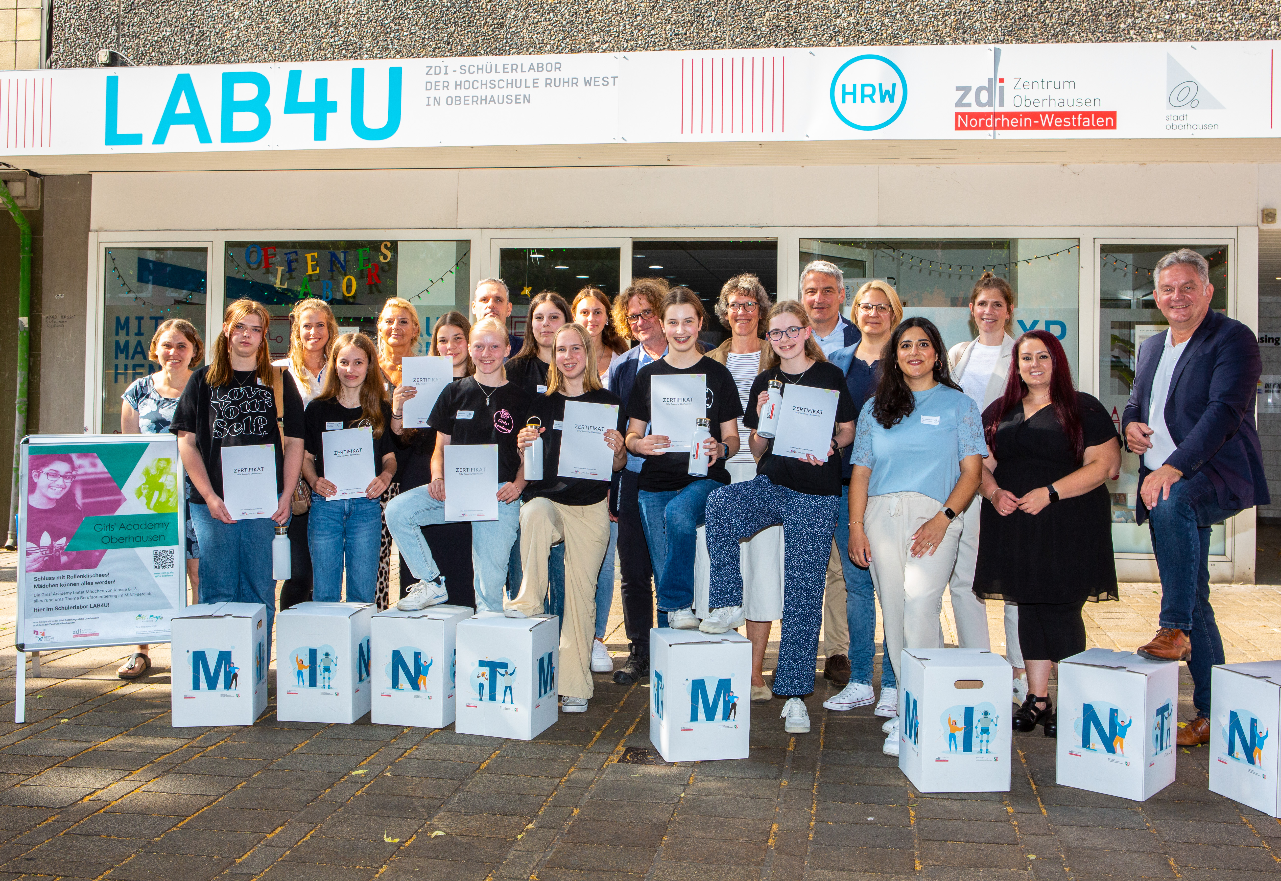 Gruppenfoto vor dem Eingang des Schülerlabors "LAB4U" der Hochschule Ruhr West in Oberhausen. Eine Gruppe junger Frauen, vermutlich Teilnehmerinnen der "Girls' Academy Oberhausen", steht gemeinsam mit Betreuenden und Dozierenden lächelnd vor dem Gebäude. Viele der Teilnehmerinnen halten ein Zertifikat und eine Trinkflasche in den Händen. Vorne stehen dekorative Würfel mit den Buchstaben „MINT“. Im Hintergrund ist ein Schild mit der Aufschrift „ZDI – Zentrum Nordrhein-Westfalen“ sowie ein Poster zur Veranstaltung zu sehen. Die Atmosphäre wirkt freundlich und feierlich.