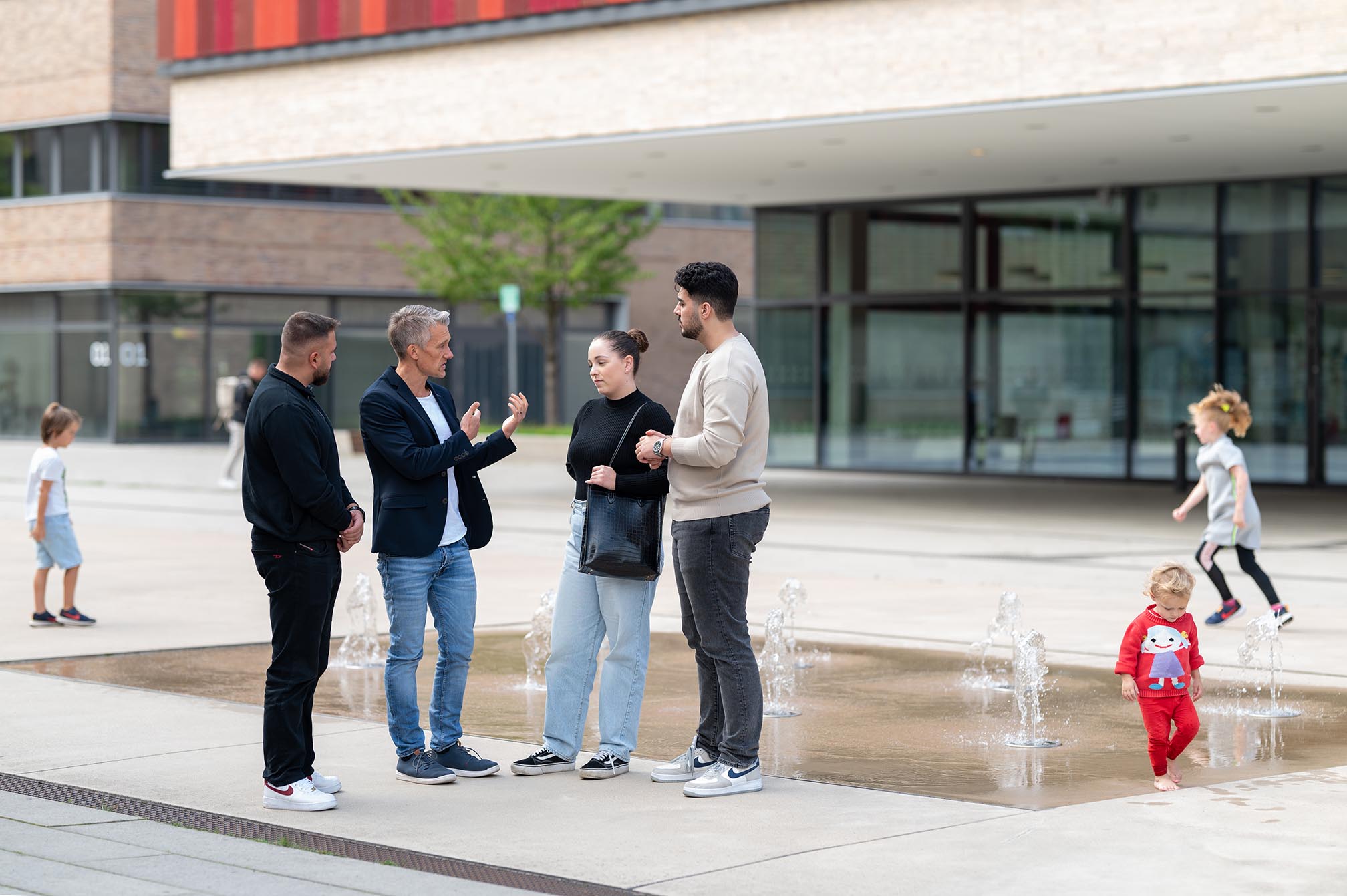 Eine Gruppe von Leuten steht vor einem der Brunnen auf dem Campus Mülheim und unterhält sich, während Kinder um den Brunnen herum spielen.