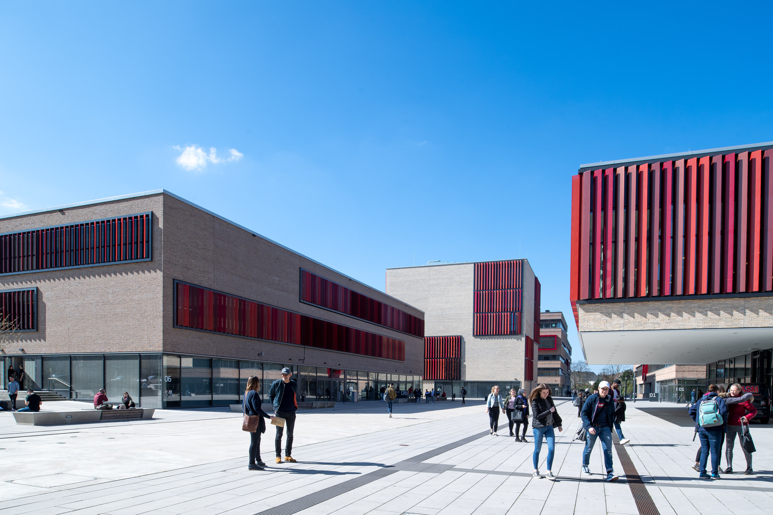 Modernes Campusgelände in Mülheim: breite Platzfläche zwischen hellen Gebäuden mit roten Lamellenfassaden, Studierende laufen und sitzen draußen bei blauem Himmel.