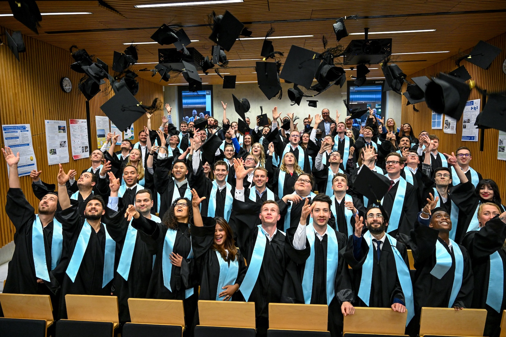 Absolvent:innen des Fachbereichs 1 (Institut Energiesysteme & Energiewirtschaft und Informatik) der Hochschule Ruhr West im Hörsaal, die gemeinsam ihre Doktorhüte in die Luft werfen.