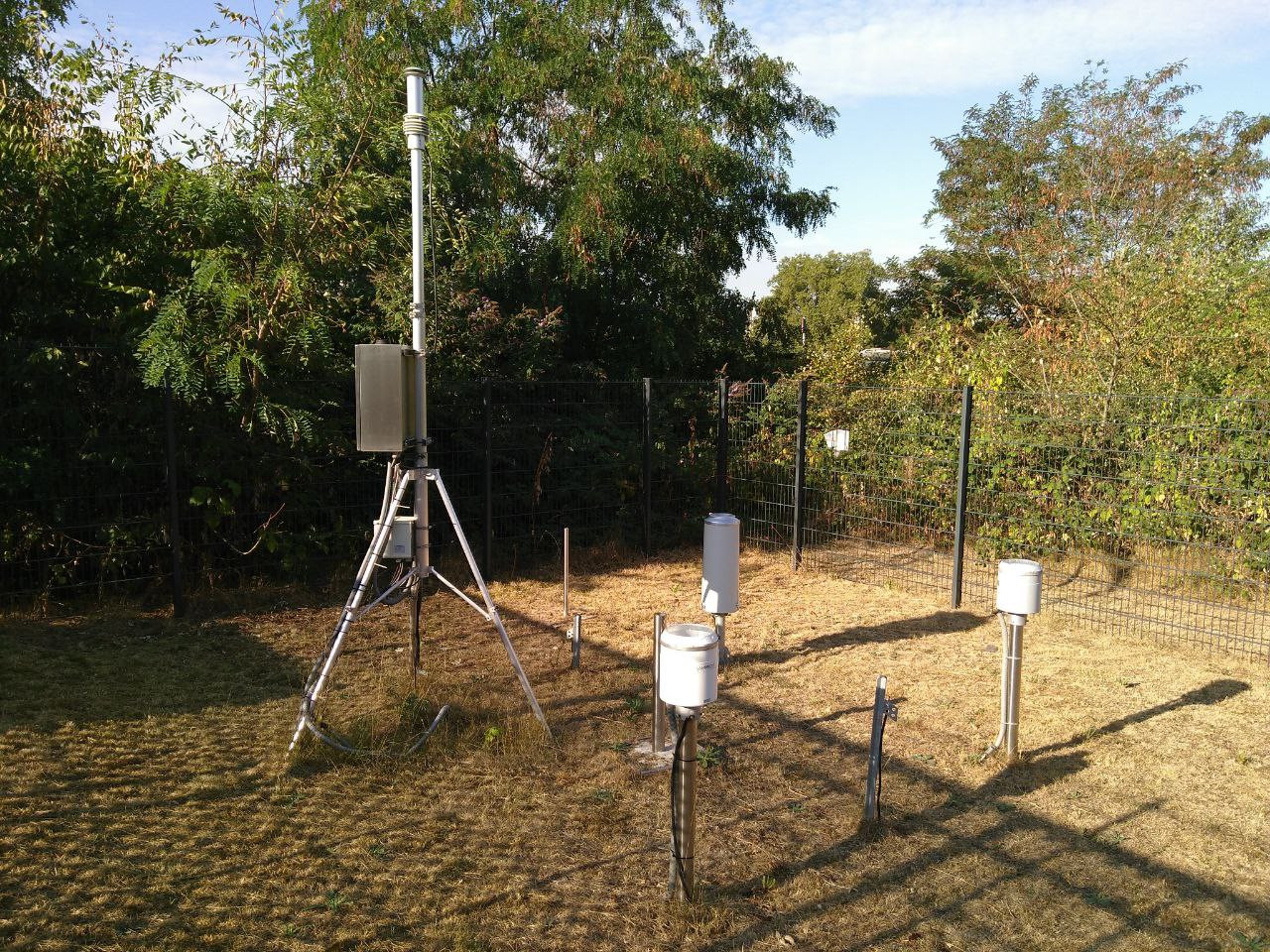 Eine Wetterstation (links) und drei Niederschlagsmesser mit unterschiedlichen Messverfahren stehen auf einem umzäunten Rasen. Hinter dem Zaun wachsen Bäume und Sträucher.