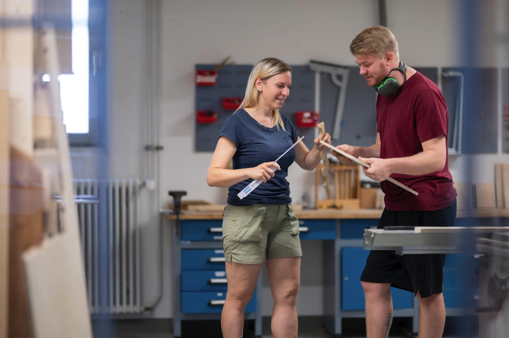 Eine Frau und ein Mann arbeiten an einer Werkbank an einem Werkstück, die Frau hält einen Zollstock