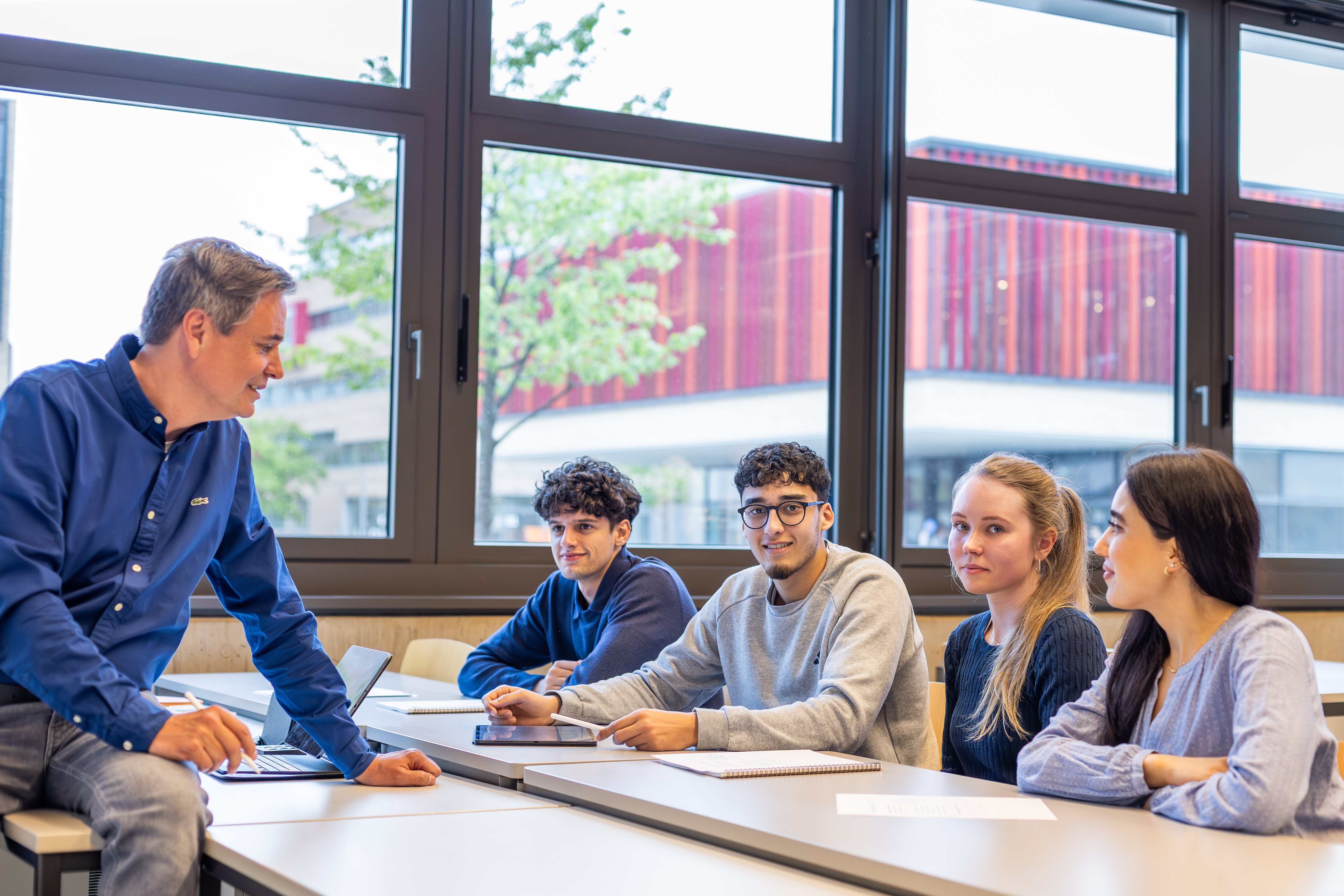 Lehrkraft spricht mit vier Studierenden im Seminarraum, an Tischen mit Unterlagen; Fenster zeigen rotes Gebäude draußen.