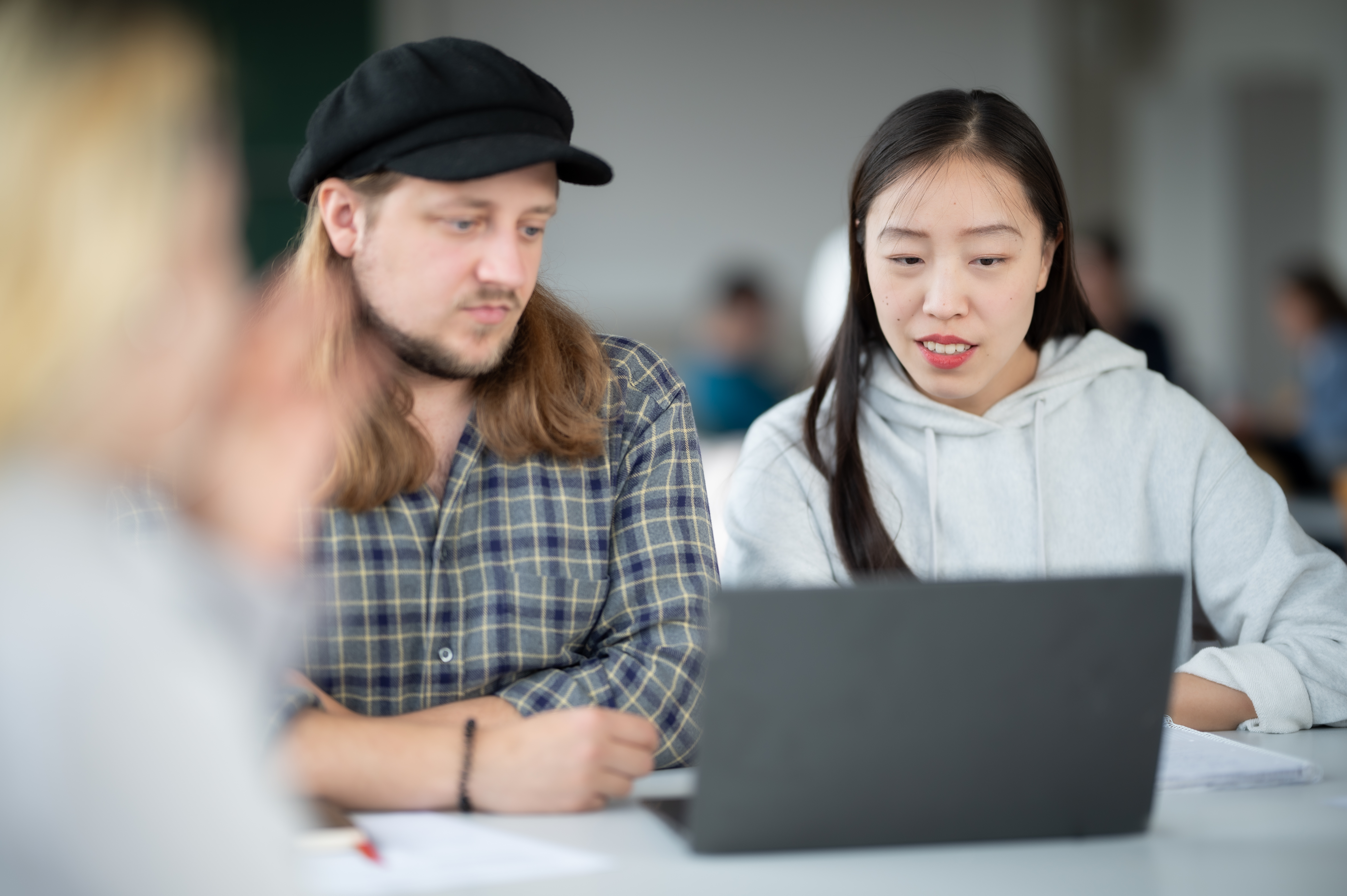 Zwei Studierende sitzen an einem Tisch und arbeiten gemeinsam an einem Laptop; eine Studentin erklärt etwas, während ein Student aufmerksam auf den Bildschirm schaut, im Hintergrund weitere unscharfe Personen in einem Seminarraum.