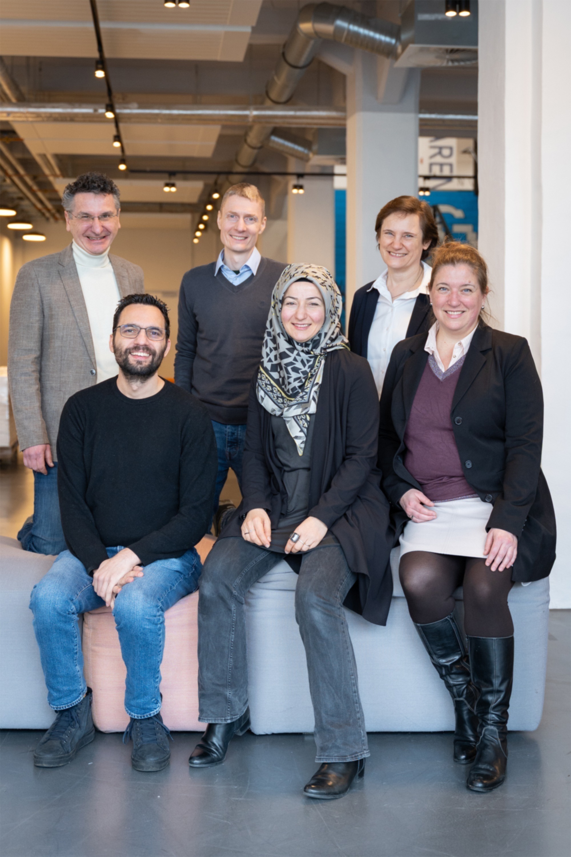 Drei Männer und drei Frauen sitzen und stehen nebeneinander in einem Raum.