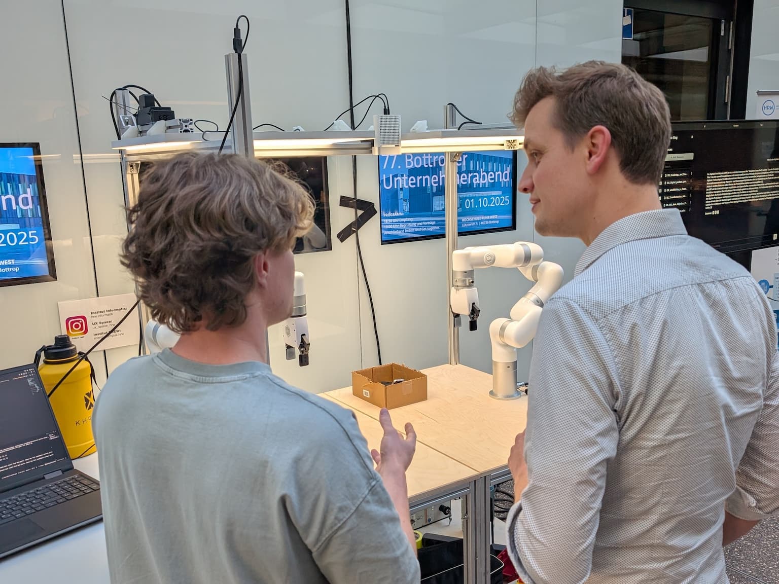 Zwei Personen stehen an einem Demonstrationsarbeitsplatz und sprechen miteinander. Auf dem Tisch befinden sich ein kollaborativer Roboterarm, ein kleiner Karton und technische Komponenten. Im Hintergrund sind Monitore mit Veranstaltungsinformationen zu sehen.