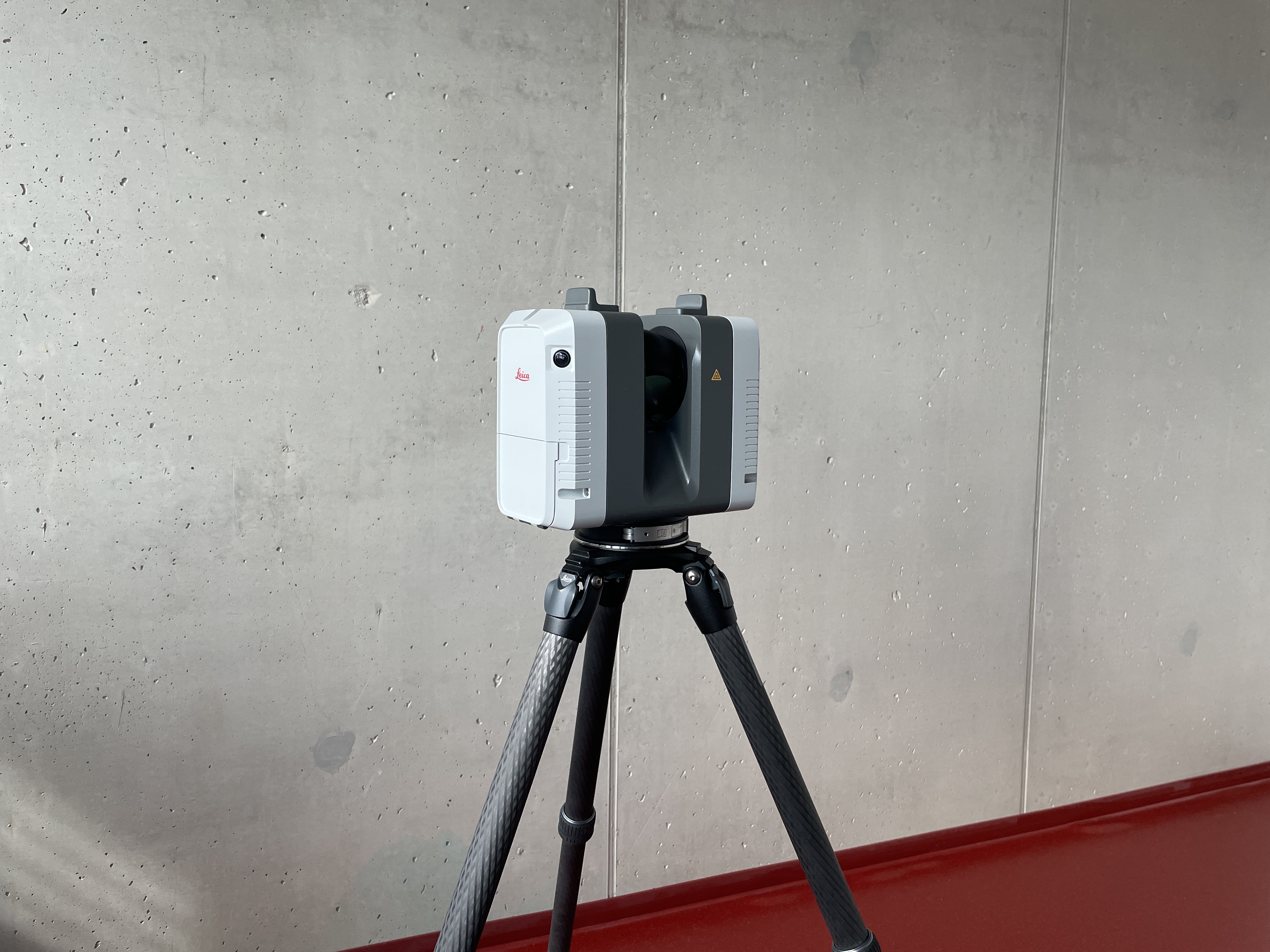 Das Bild zeigt einen Laserscanner vor einer grauen Wand und rotem Boden