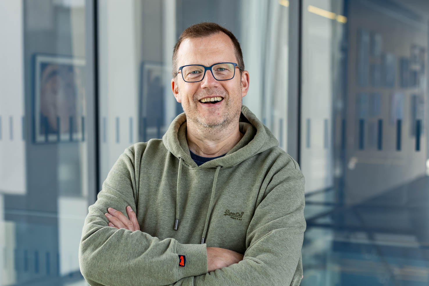 Lächelnder Mann mit Brille im grünen Hoodie, Arme verschränkt, steht in einem modernen verglasten Flur.