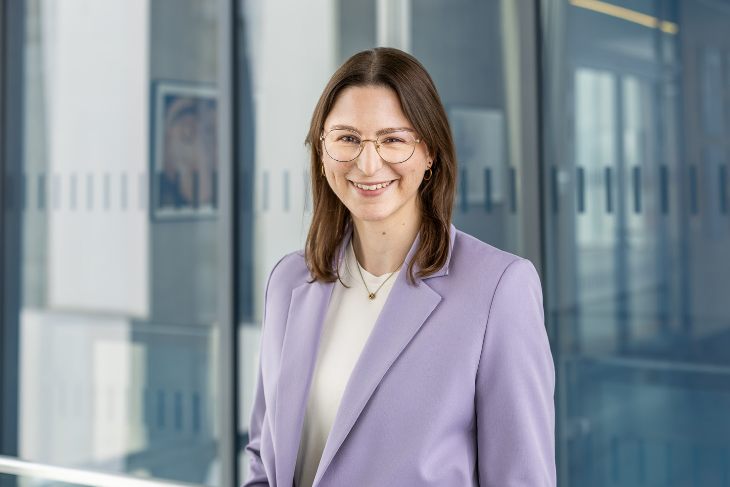 Lächelnde Person mit Brille im hellvioletten Blazer vor moderner Glaswand, professionelles Büro-Porträt.