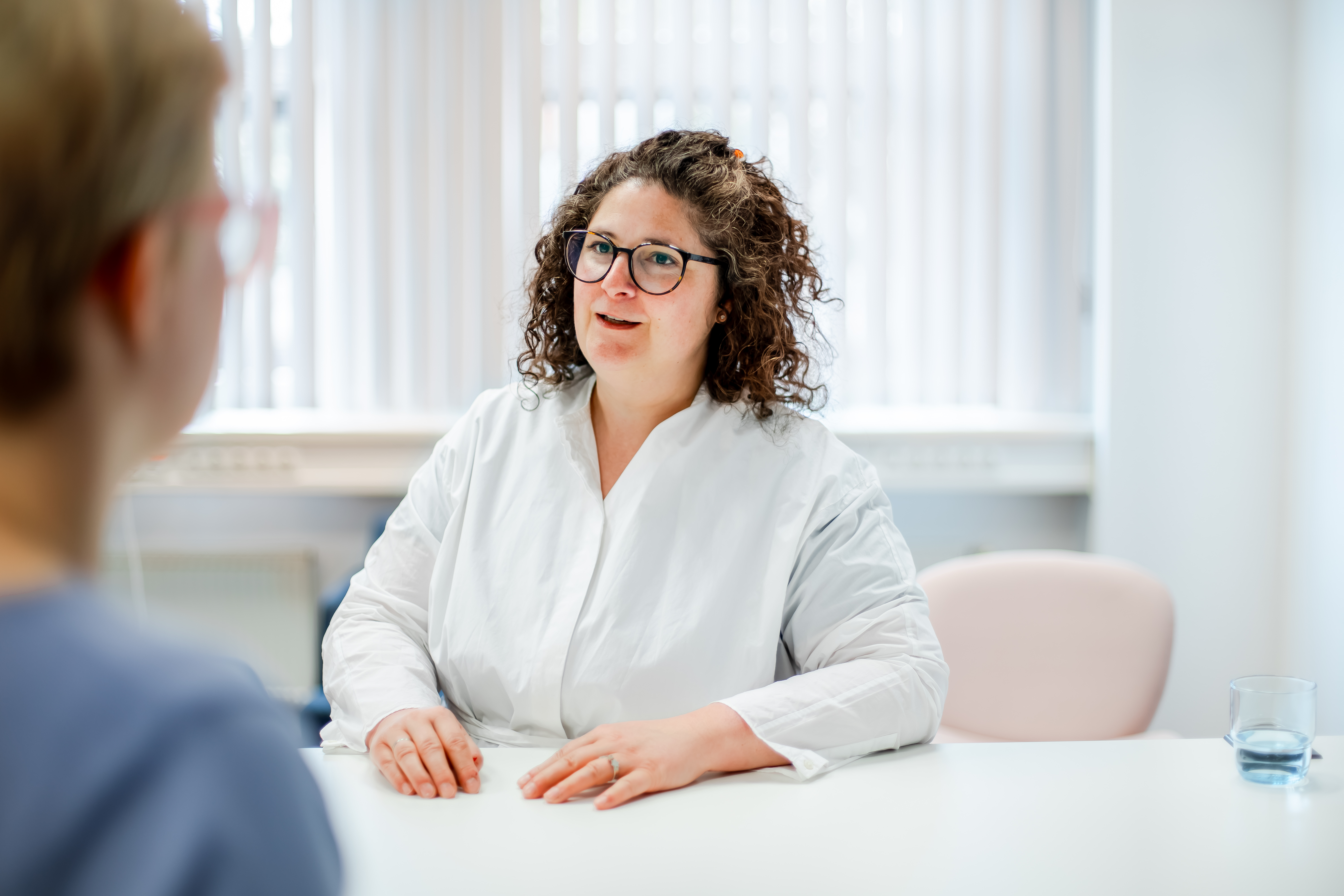 Auf dem Foto ist eine Frau mit dunklen Haaren und Brille zu erkennen. Sie spricht mit einer Person, die ihr gegenüber sitzt.