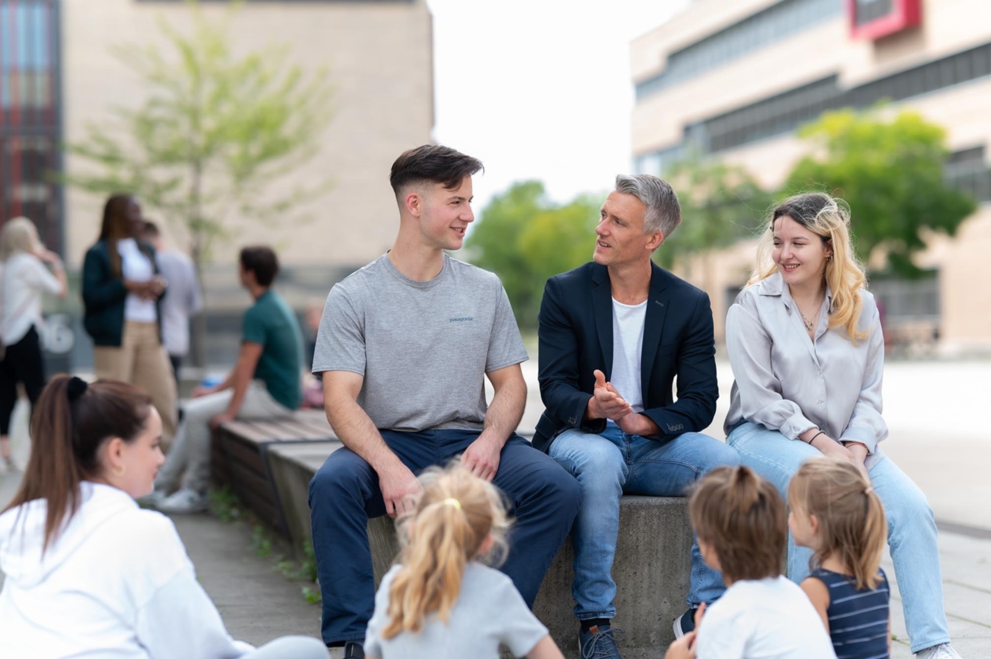 Ein Mann sitzt zusammen mit Studierenden und spricht mit Kindern auf dem Campus Mülheim.