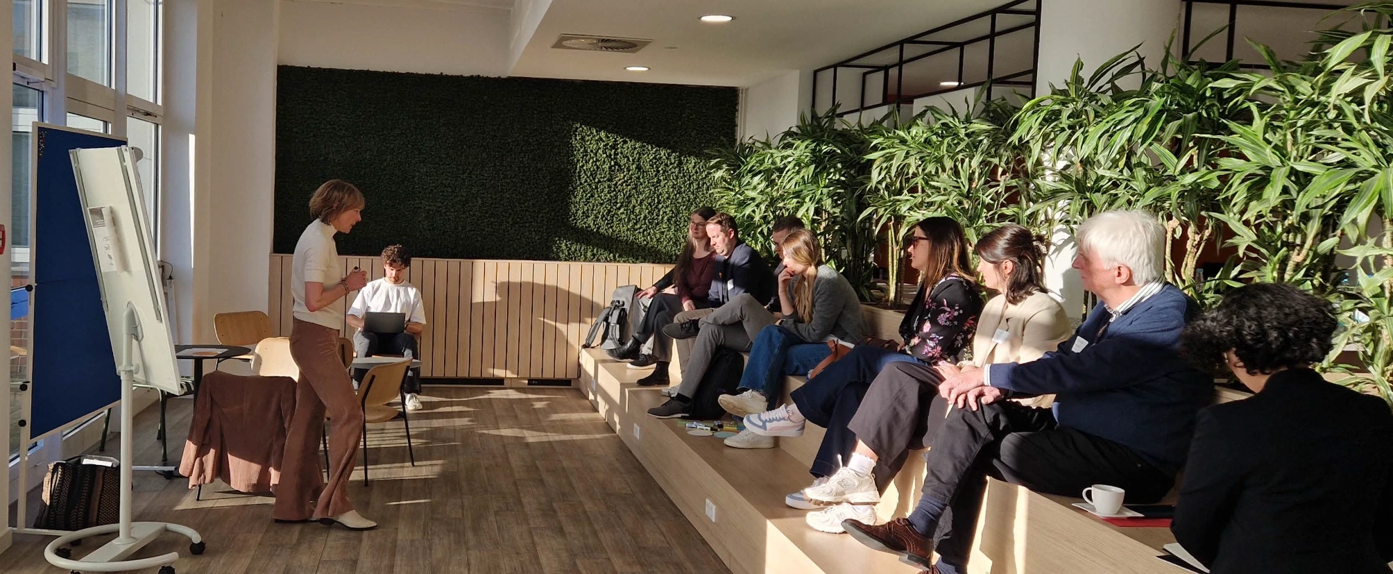 Eine Person steht vor einem Flipchart und spricht zu einer Gruppe von Teilnehmenden, die in einem hellen Raum mit vielen Pflanzen auf einer Sitzstufe sitzen und zuh&ouml;ren.
