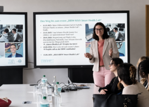 KKS Smart Health Lab / Carole Leguy: Eine Frau steht zwischen Bildschirmen und einer Gruppe junger Menschen, die an einem Tisch sitzen.