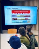 Mehrere Schüler:innen sitzen an einem Tisch und schauen auf einen großen Bildschirm, auf dem eine Präsentationsfolie zu ‚Category Management‘ mit Regalgrafiken aus dem Supermarkt zu sehen ist.