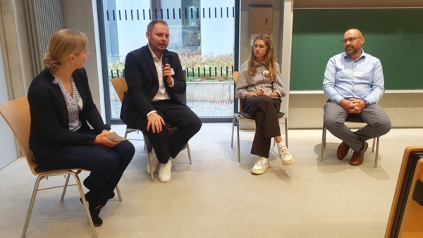 Vier Personen sitzen in einer Podiumsdiskussion; eine Person spricht ins Mikrofon. Innenraum mit Tafel und gro&szlig;en Fenstern im Hintergrund.