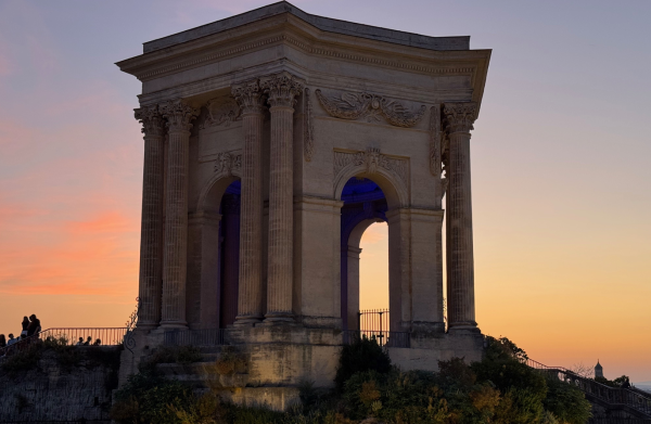Ein klassizistischer Pavillon mit Säulen im Gegenlicht eines farbenfrohen Sonnenuntergangs, Menschen im Vordergrund.